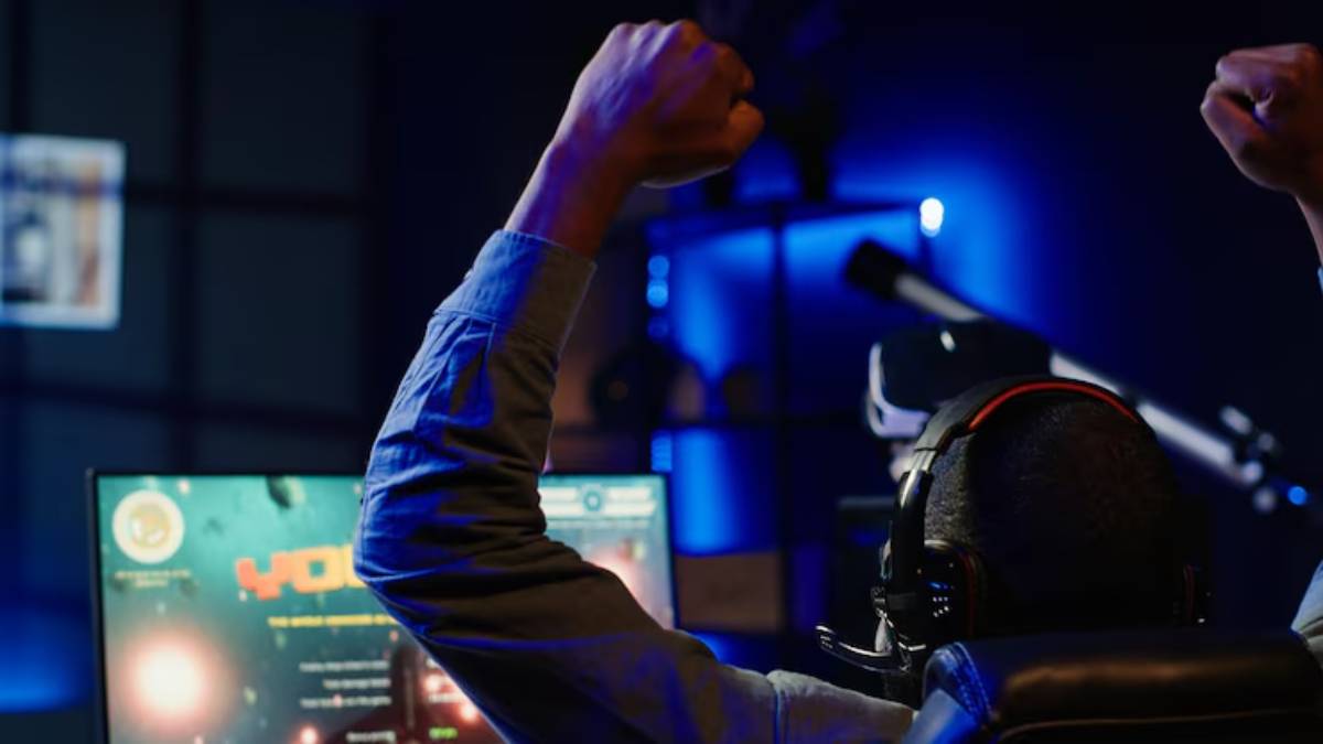 A gamer celebrates a victory, raising both fists in triumph, while wearing a headset in a dimly lit, modern gaming setup. 