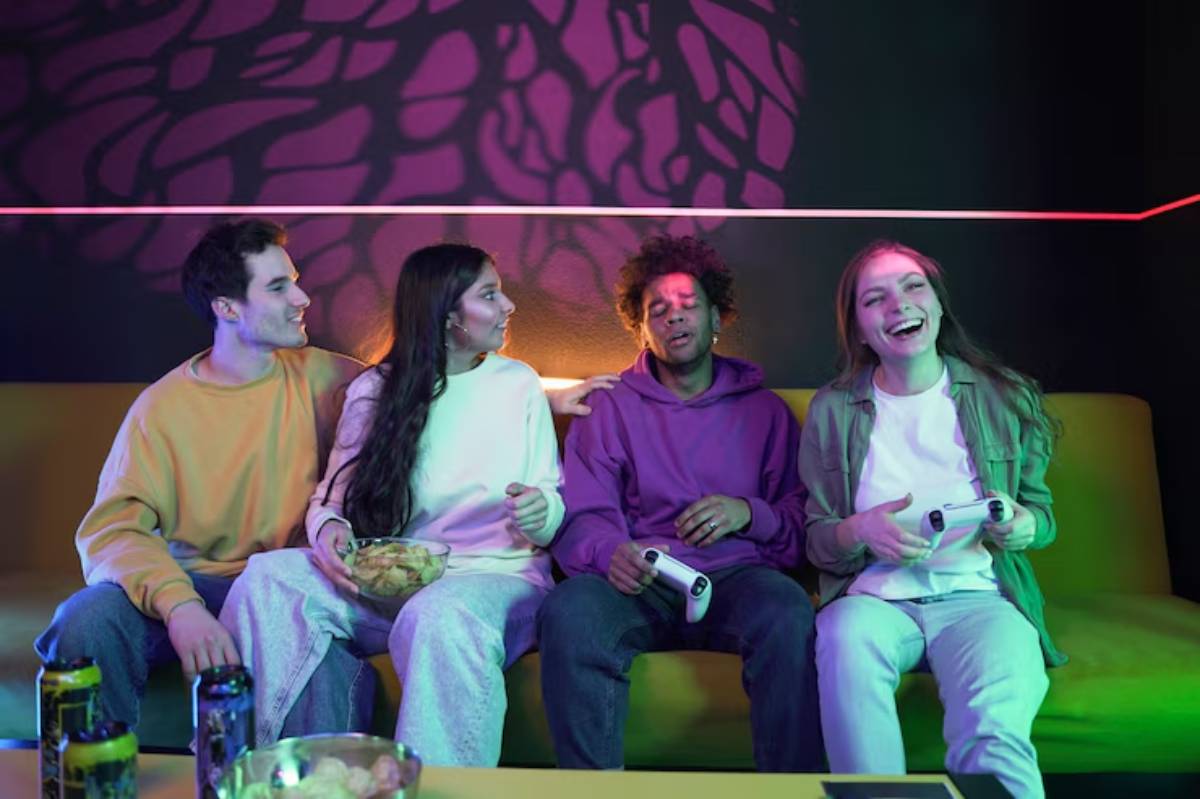 A group of four friends sits together on a couch, playing video games, surrounded by colorful ambient lighting and snacks.
