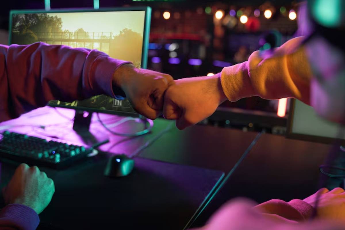 Two hands perform a fist bump over a desk with gaming equipment, illuminated by colorful lights in a vibrant gaming environment.