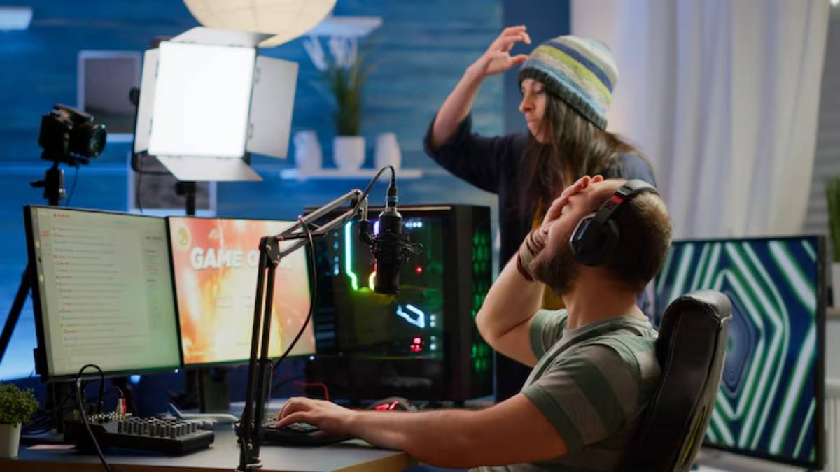 A gamer sits at a desk with multiple monitors, headphones on, showing frustration while a person stands behind them, gesturing playfully.