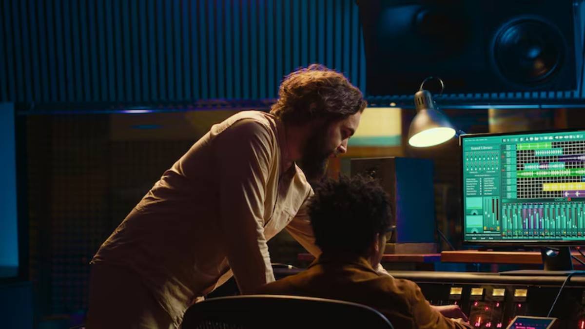 Two individuals are collaborating in a music studio, focused on a computer screen displaying sound editing software and audio tracks.