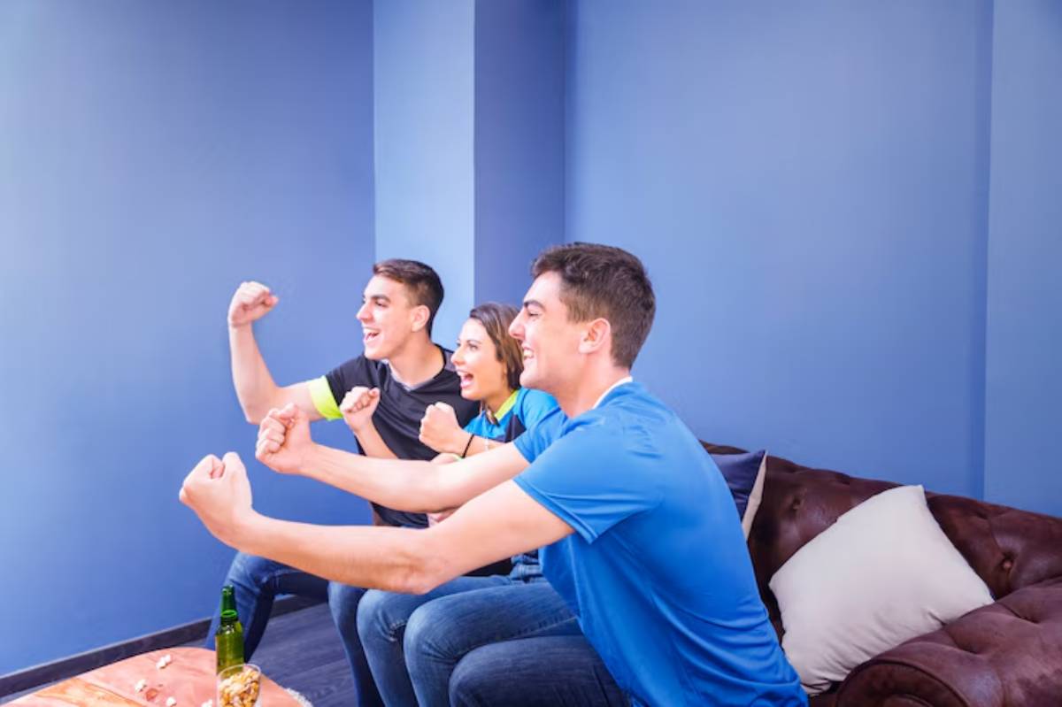Three people cheer enthusiastically while watching a sports event, seated on a brown couch in a blue-walled room, snacks and drinks nearby.