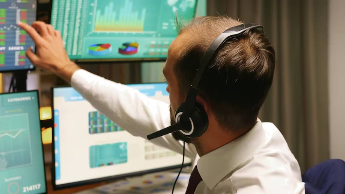 A man in a suit and headset analyzes data on multiple computer screens filled with charts and graphs in a modern office setting.