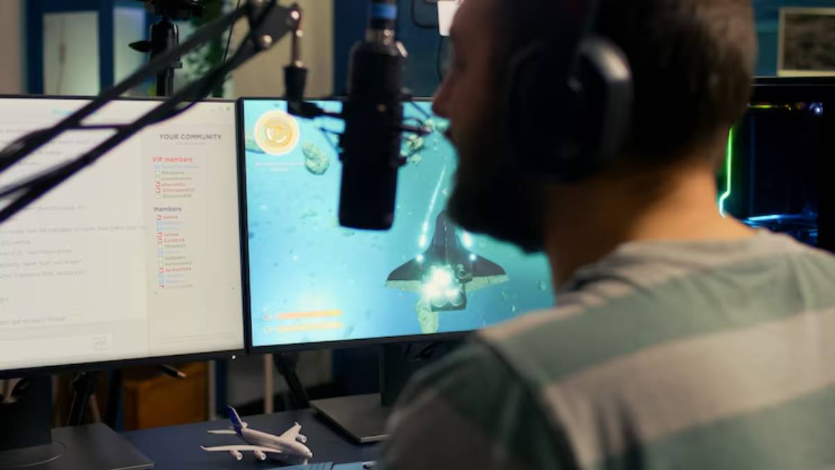 A person plays a video game featuring an underwater scene on dual monitors, with a microphone and a small airplane model in view.