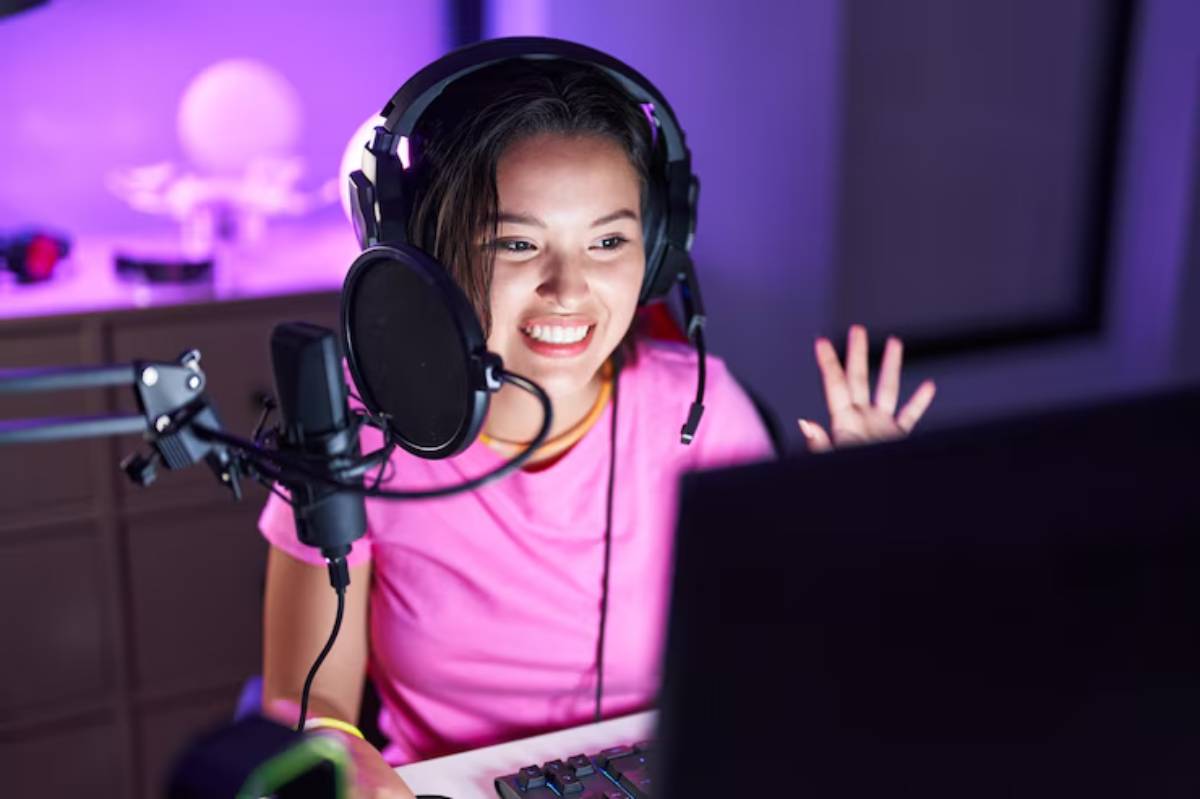 A person wearing headphones sits in front of a computer, gesturing while speaking into a microphone, surrounded by purple lighting.