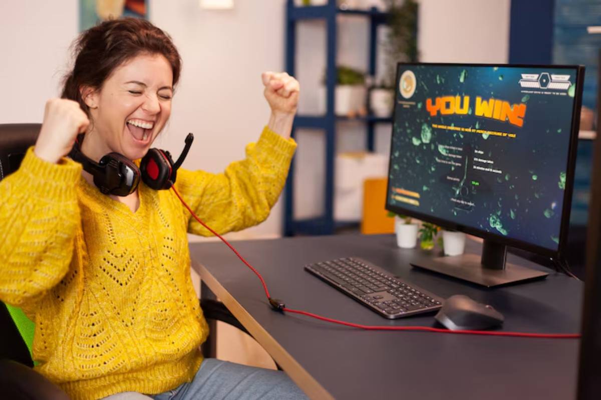 A person in a yellow sweater celebrates victory in an online game, wearing headphones, with a computer displaying 