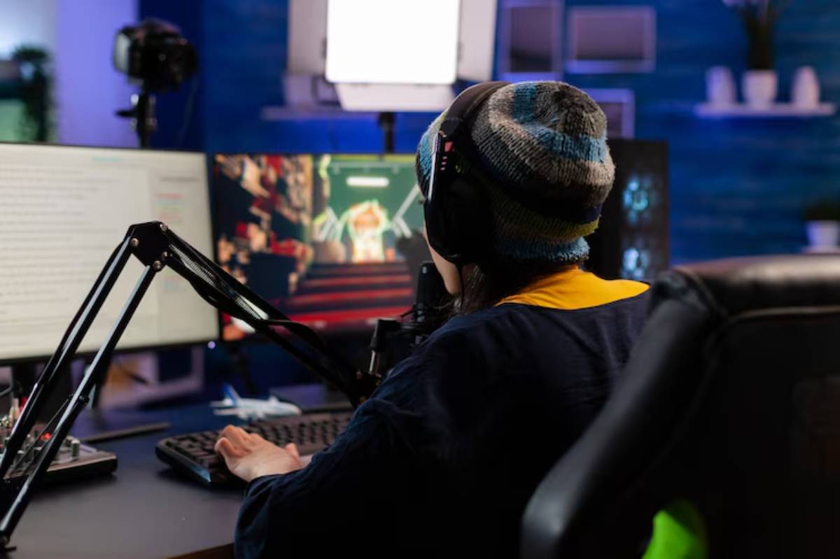 A gamer wearing headphones and a beanie focuses on dual monitors, one displaying a vibrant game scene, amidst a colorful gaming setup.