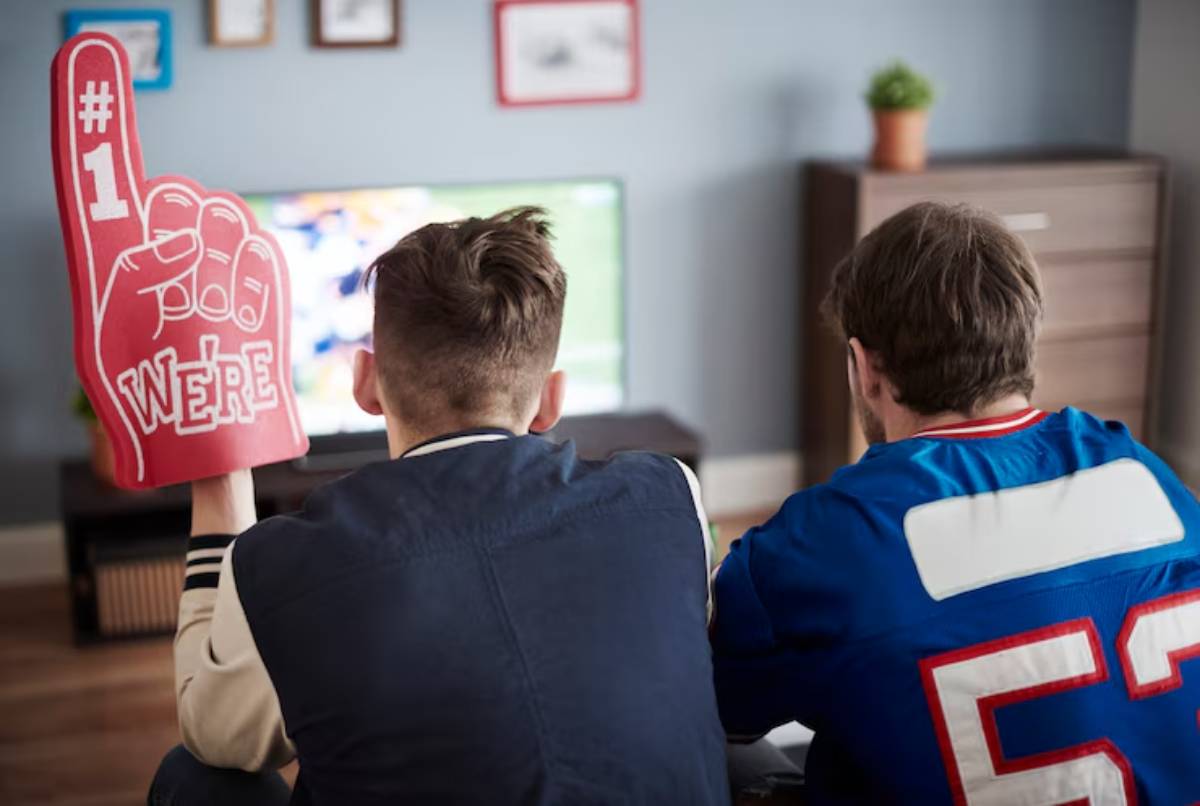 Two friends watch a sports game on TV, one holding a #1 We're foam finger, both wearing team apparel, creating a lively atmosphere.