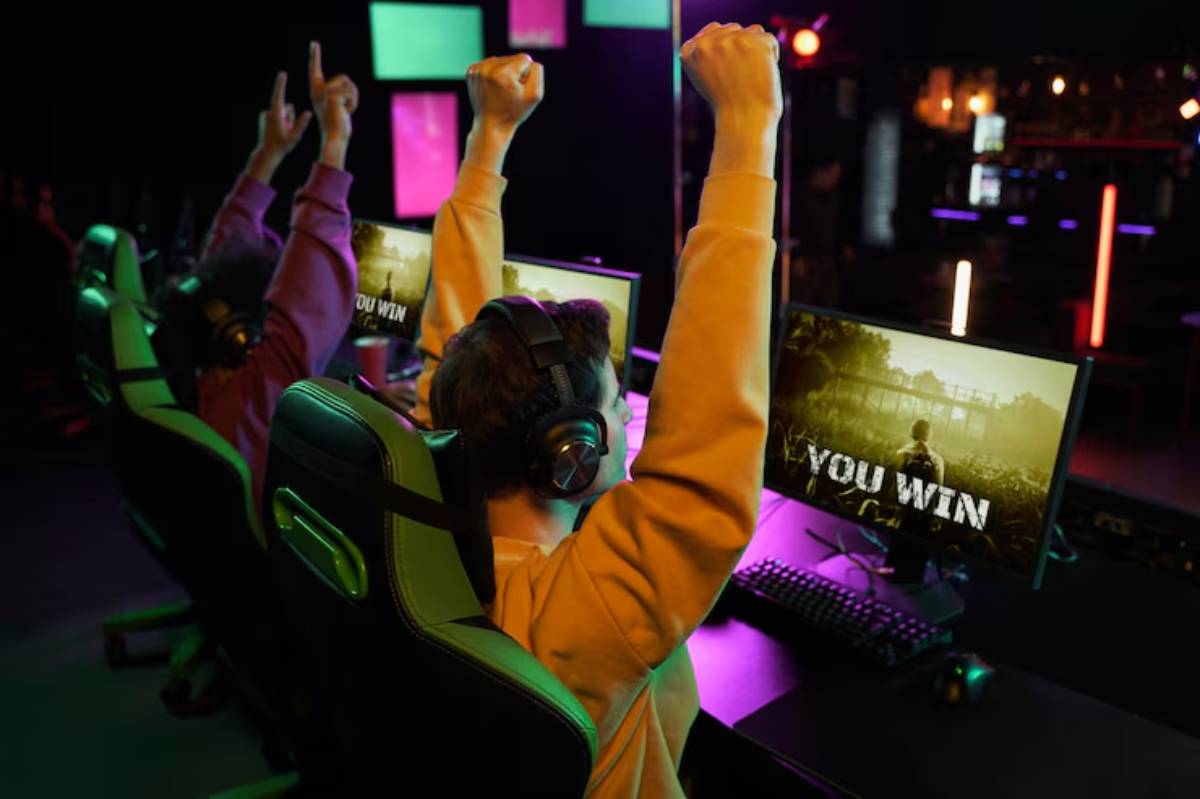 Two gamers celebrate victory in an esports setting, raising their arms in excitement as "YOU WIN" appears on their screens.