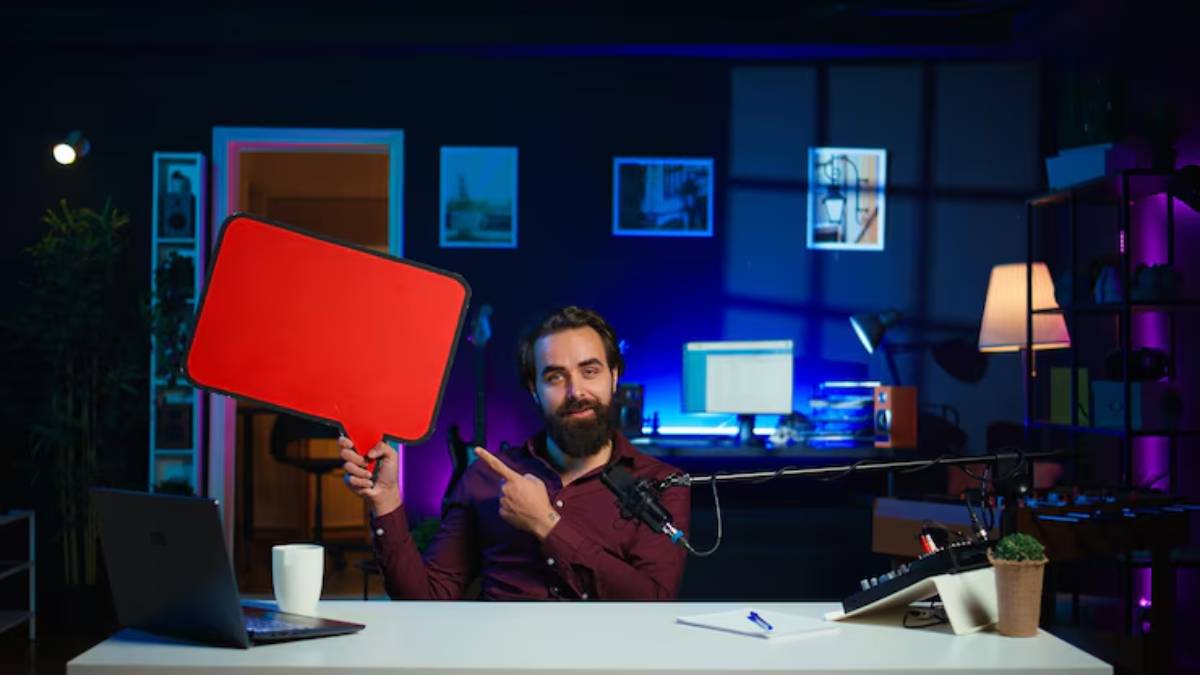 A person sits at a desk in a modern workspace, holding a large red speech bubble sign while gesturing towards it, surrounded by tech gadgets.