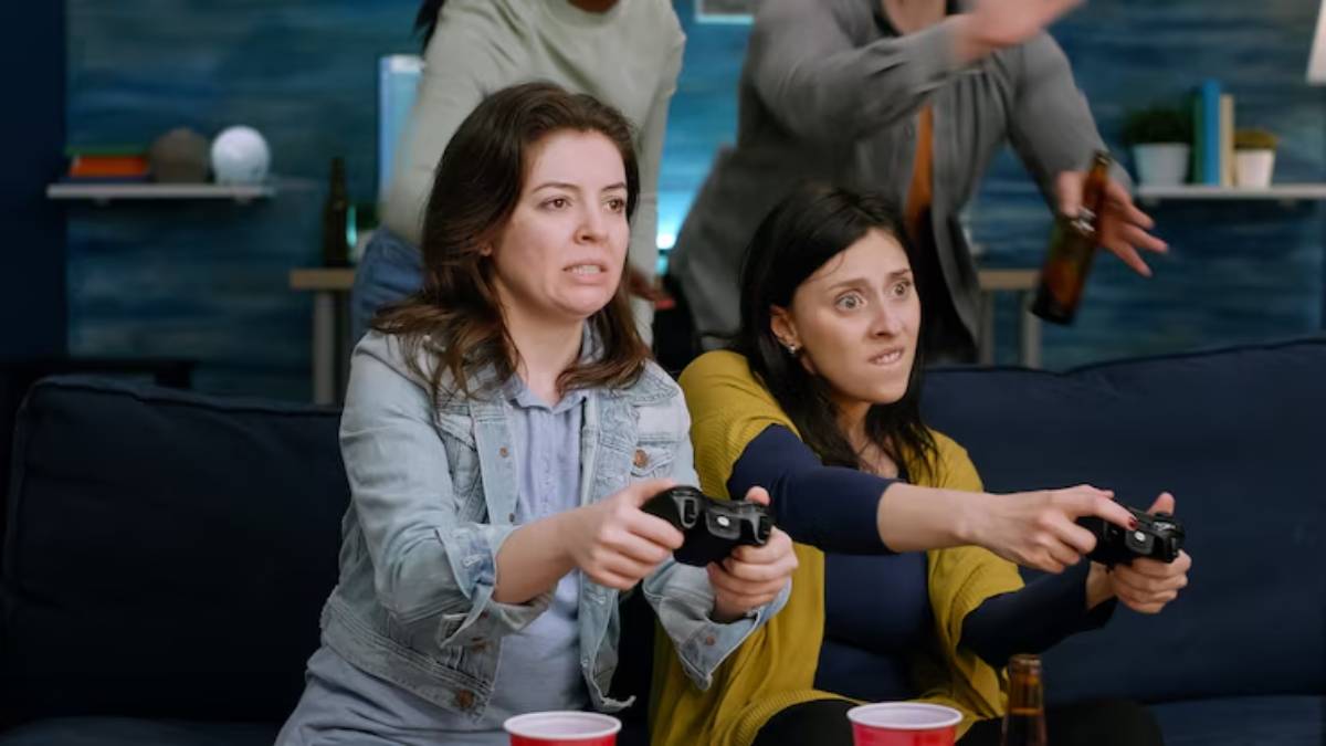 Two women are sitting on a couch, intensely focused on playing video games, while others cheer in the background. Red cups are on the table.