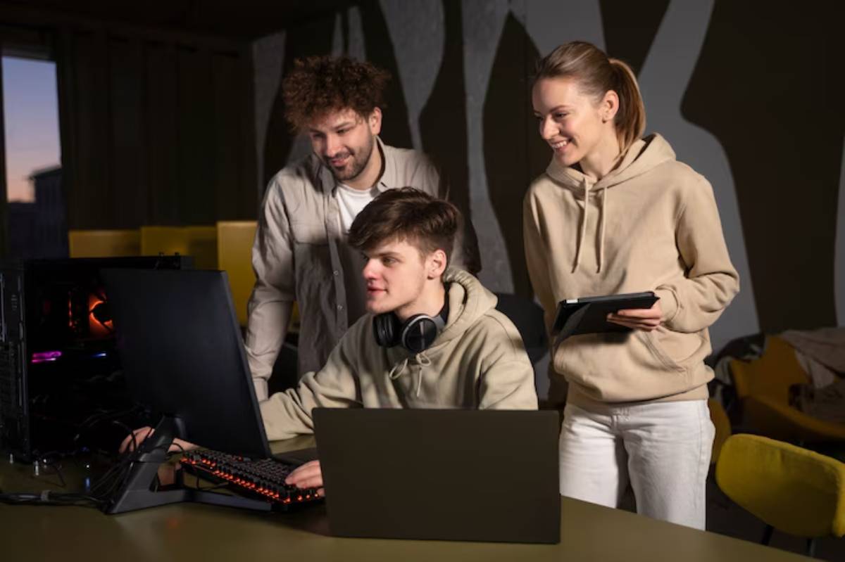 Three individuals in beige hoodies collaborate around computers, engaged in a gaming or coding session in a cozy, modern setting.