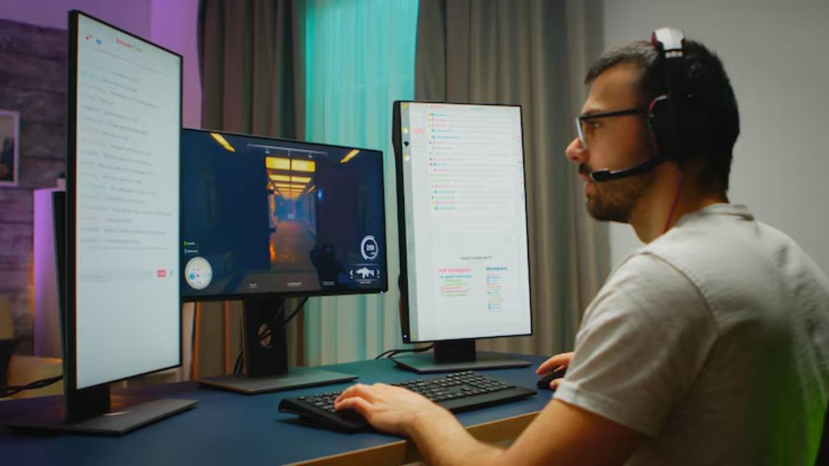 A gamer intensely focused on dual monitors, playing a game and viewing a chat interface, wearing headphones in a modern setup.