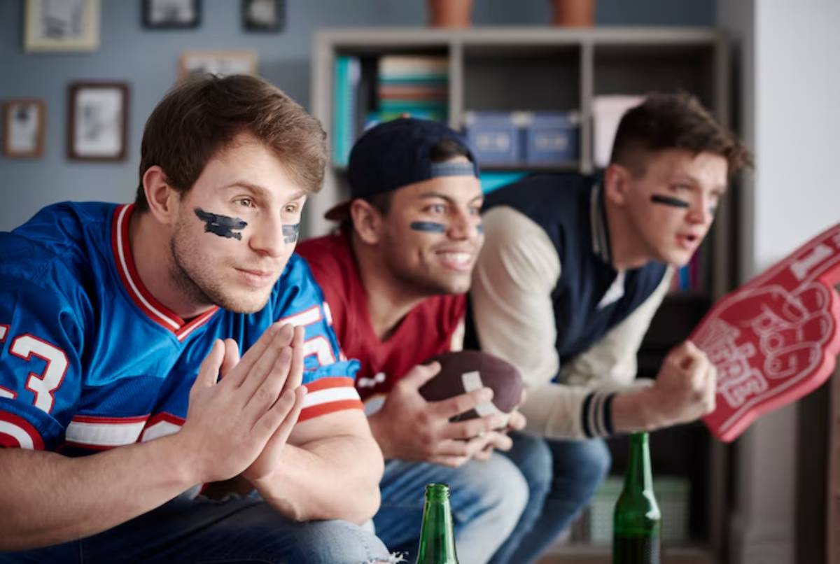 Three men in sports jerseys anxiously watching a game, holding a football and a foam finger, with drinks on a table.