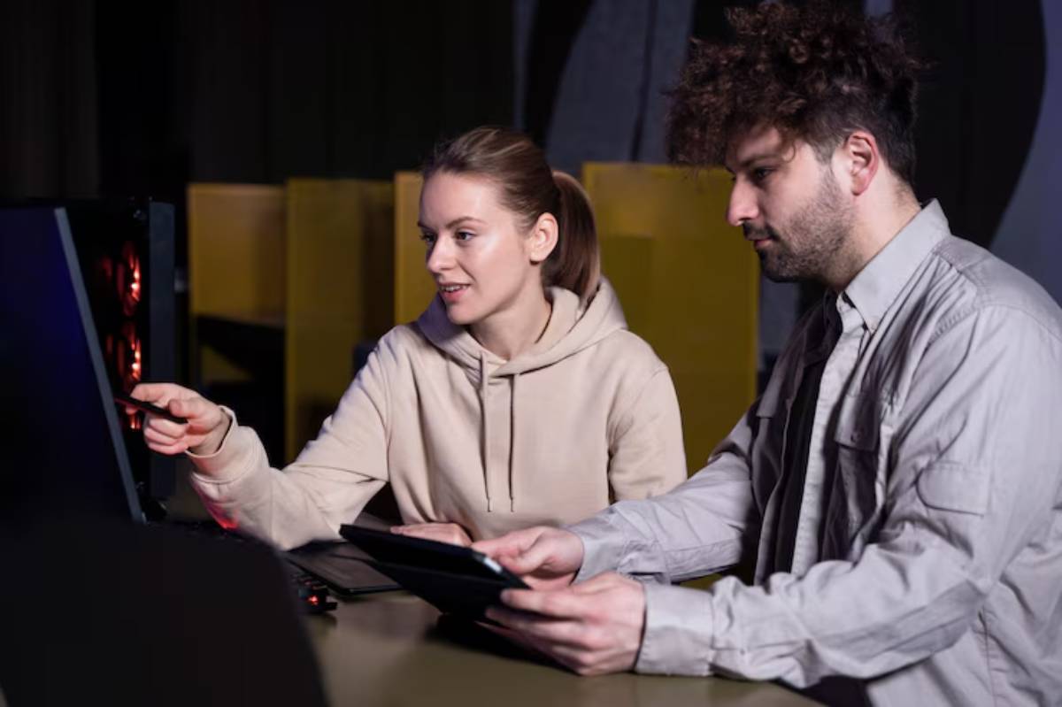 A woman in a beige hoodie points at a computer screen while a man holds a tablet, both engaged in a discussion.