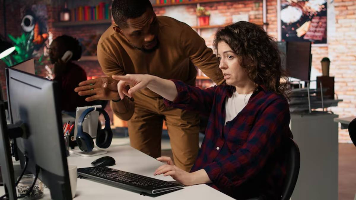 A person at a computer demonstrates something to another individual in a tech-focused workspace, with colorful decor in the background.