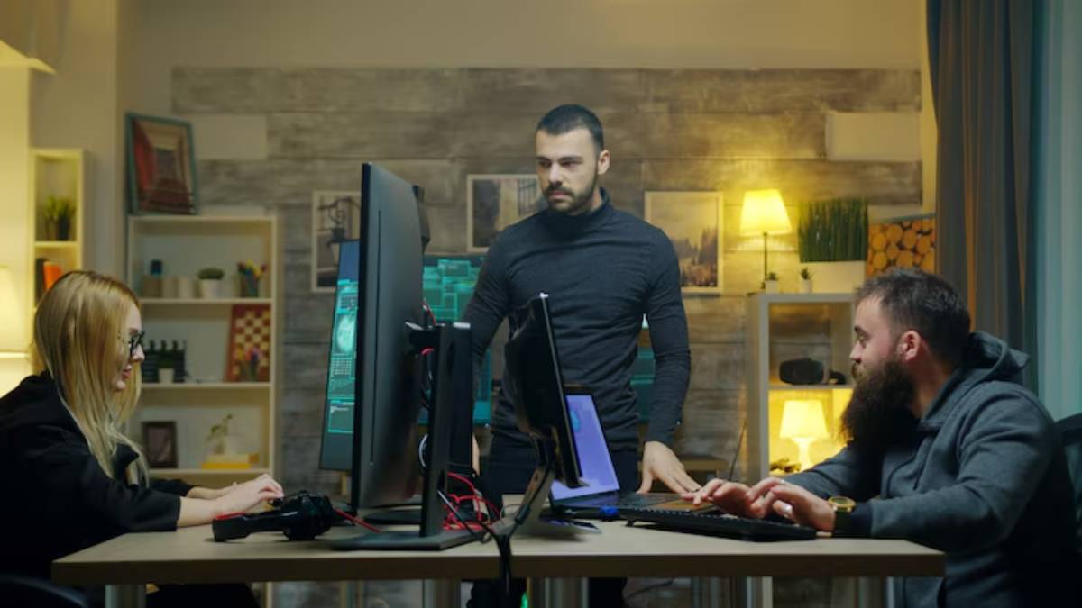 A group of three people collaborates around a table with multiple computer screens and gaming equipment in a cozy, modern workspace.