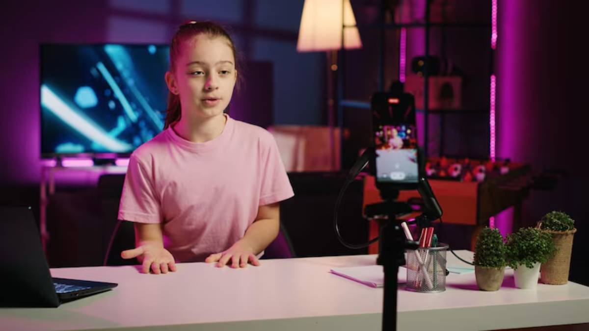 A person in a pink T-shirt gestures while sitting at a desk, with a laptop and potted plants in a stylish, illuminated room.
