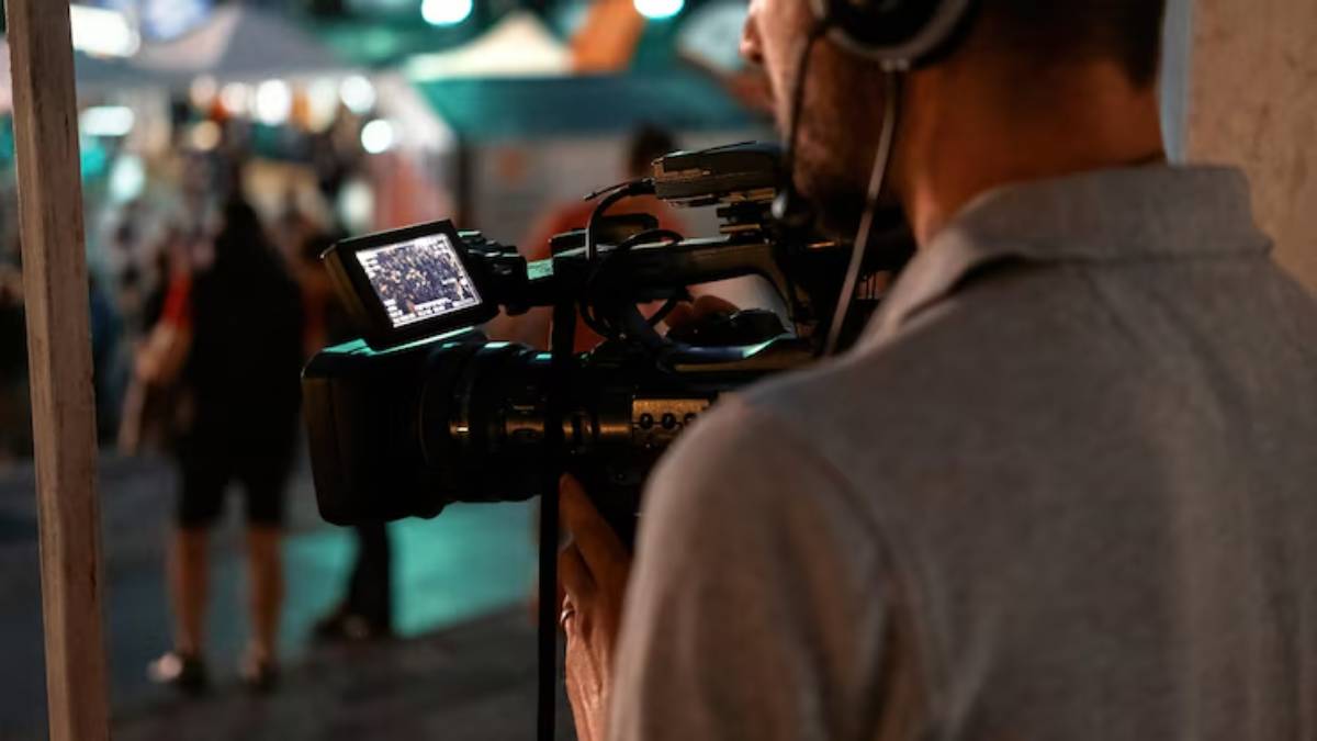 A videographer captures footage at an outdoor event, with a camcorder displaying live action and festival lights in the background.