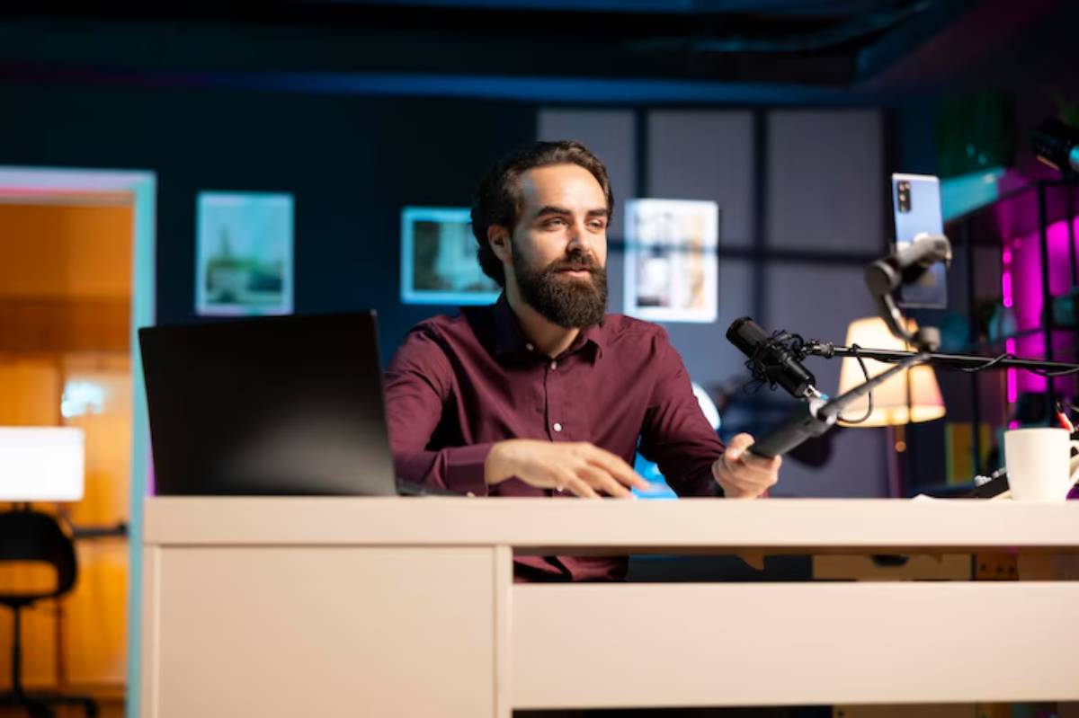 A person in a maroon shirt gestures while seated at a desk with a laptop and microphone, surrounded by colorful ambient lighting.