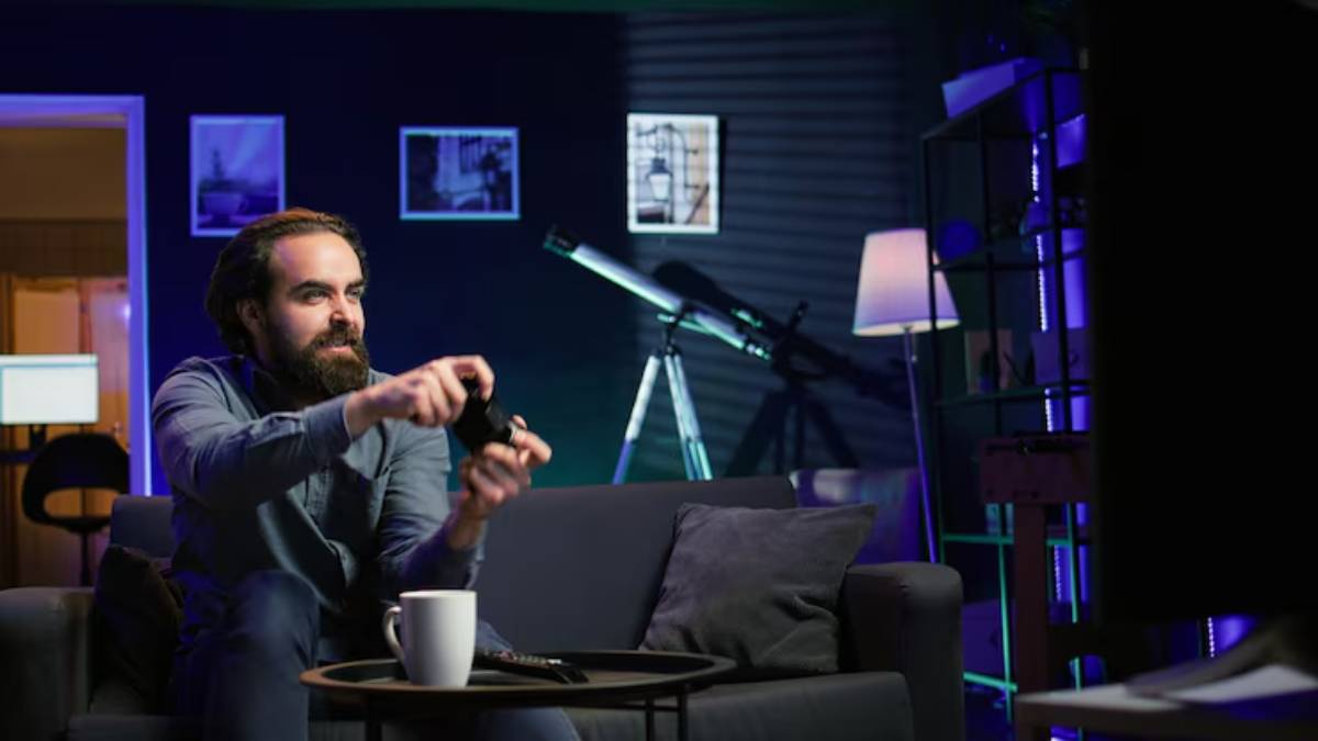 A person sits on a couch in a cozy room, holding a gaming controller with a telescope and soft lighting in the background.