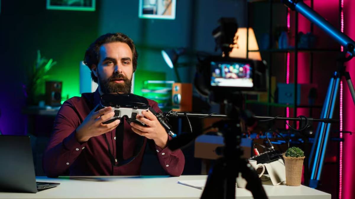 A person holds a device in a dimly lit room, surrounded by colorful lights and a camera focused on them, with a laptop and plant nearby.