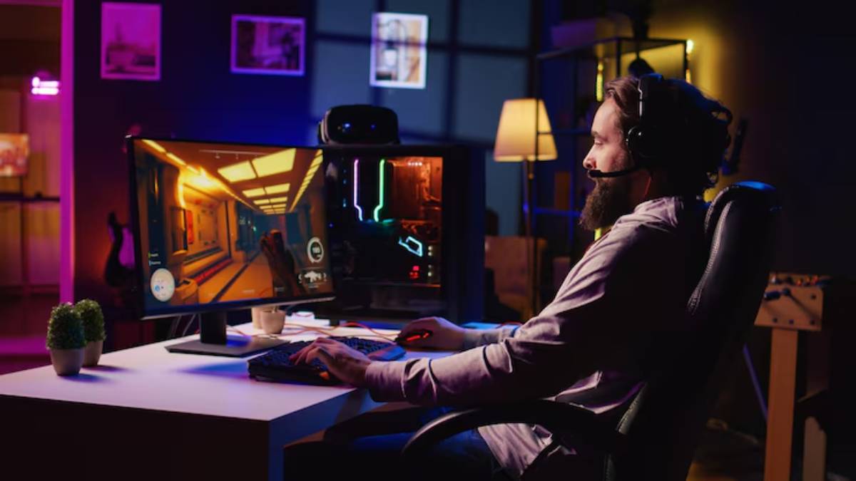 A gamer sits at a desk, focused on dual monitors displaying a vivid video game in a dimly lit, colorful room.