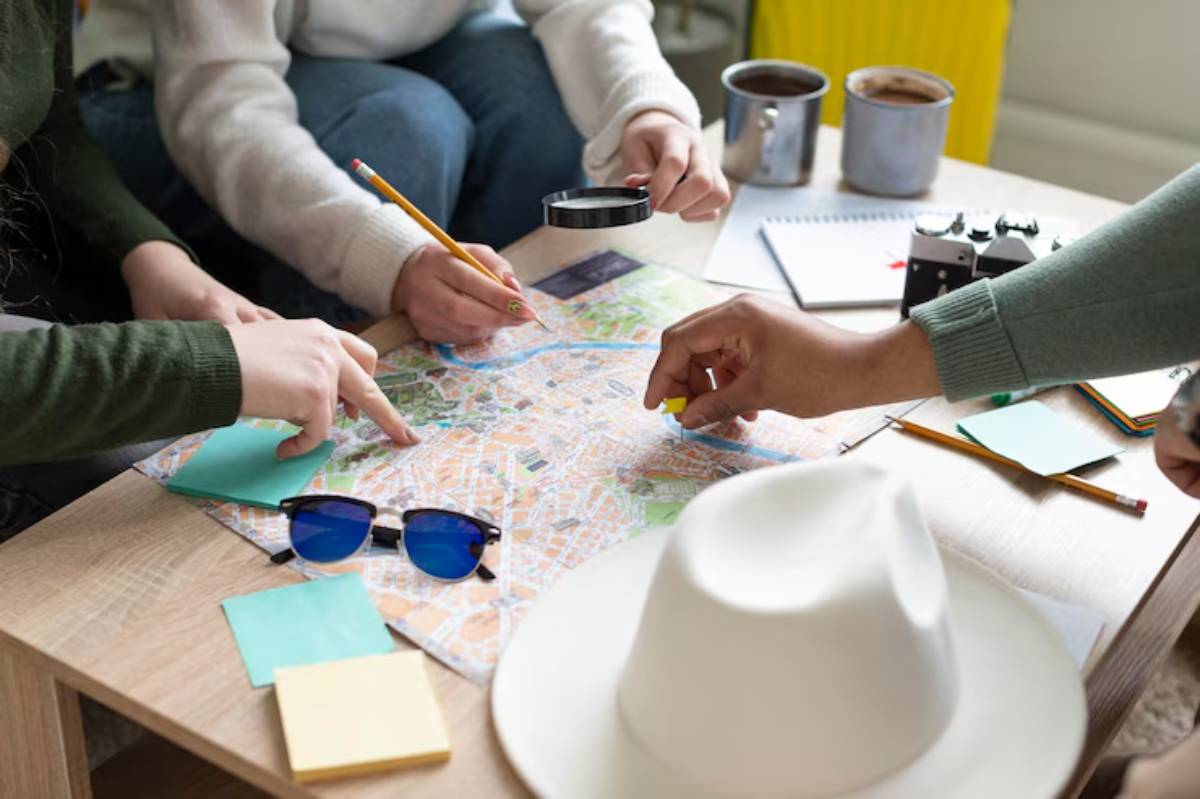 Four people study a city map on a table, with a magnifying glass, pencils, sticky notes, sunglasses, and coffee cups nearby.