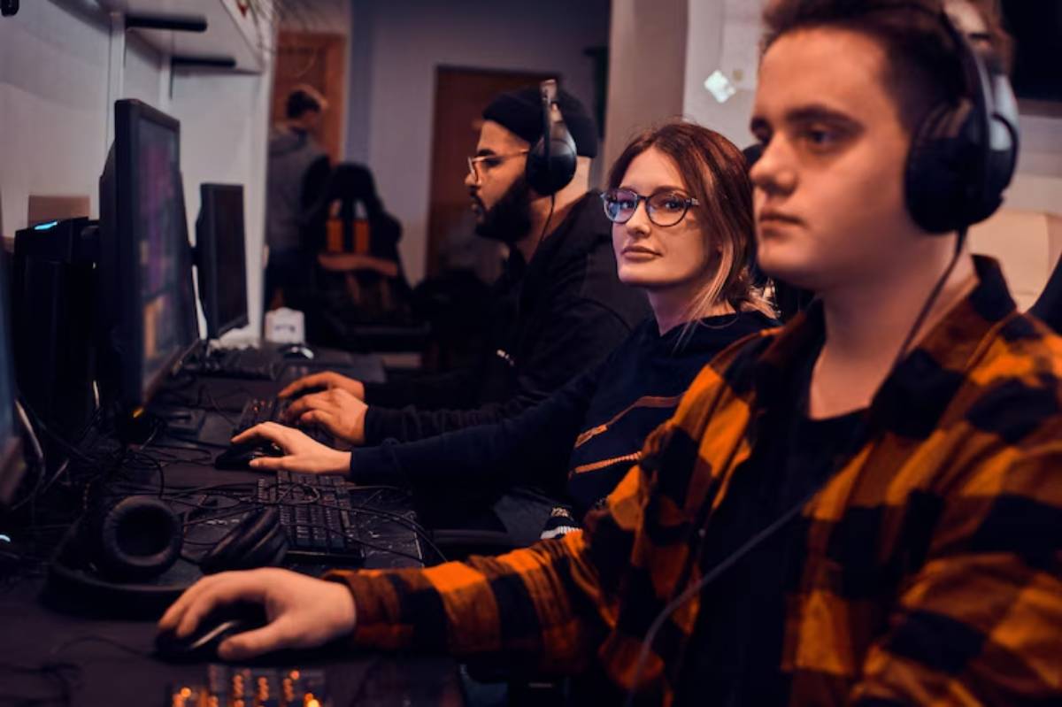 Four individuals sit at a gaming station, absorbed in their screens, wearing headphones, with keyboards and mice in front of them.