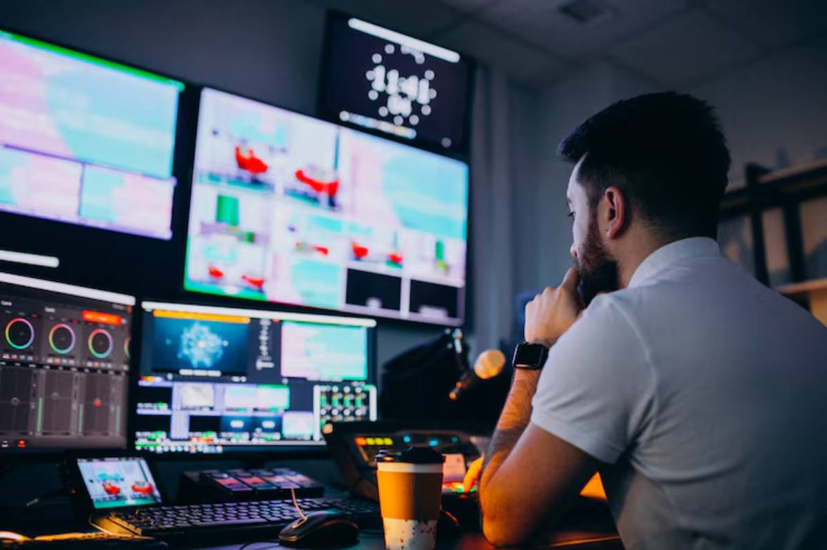 A focused man in a control room monitors multiple screens displaying colorful graphics and video feeds, with a coffee cup nearby.