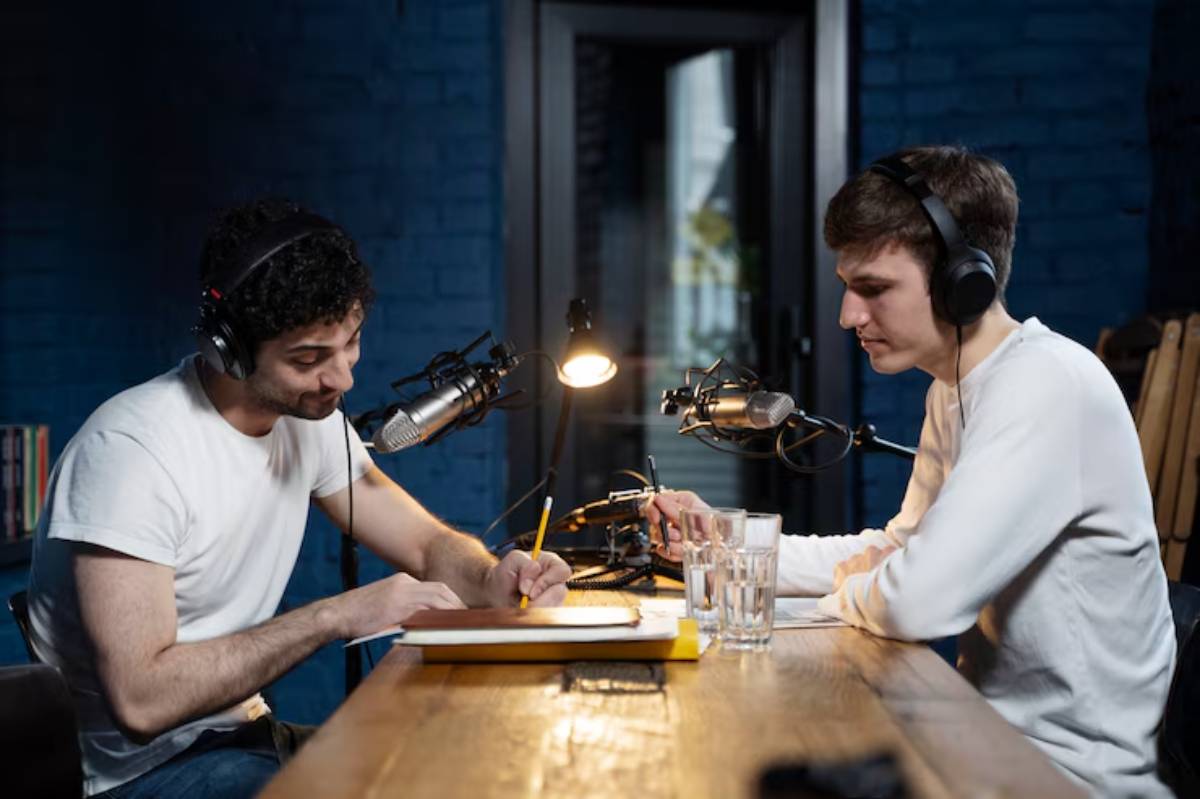 Two individuals in headphones sit at a wooden table with microphones, engaged in a podcast discussion, amid a blue brick backdrop. 