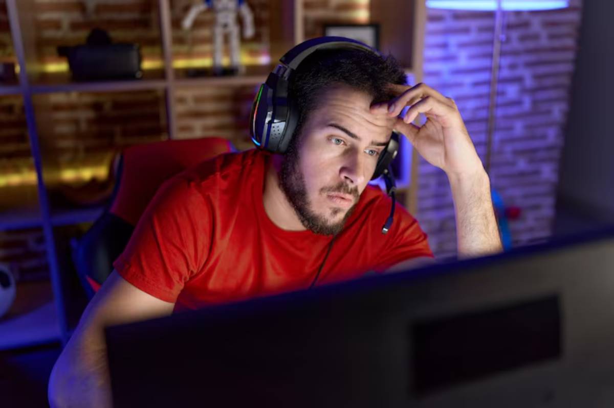 A person in a red shirt and headphones sits in front of a computer, looking stressed in a dimly lit gaming room.
