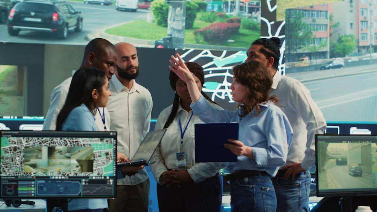A diverse group discusses surveillance data in a high-tech monitoring room, with computer screens displaying live traffic and city maps.