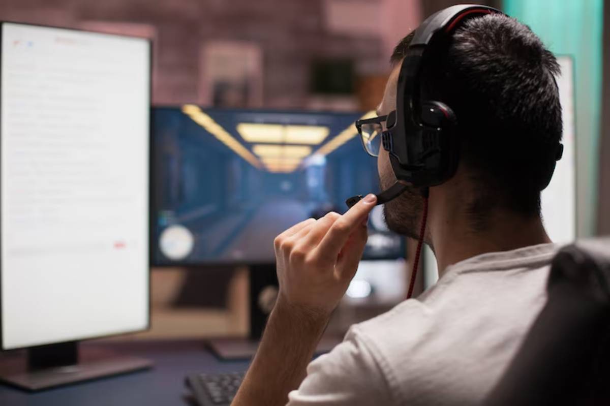 A gamer wearing a headset sits at a desk, focused on dual monitors displaying gameplay and a chat window.
