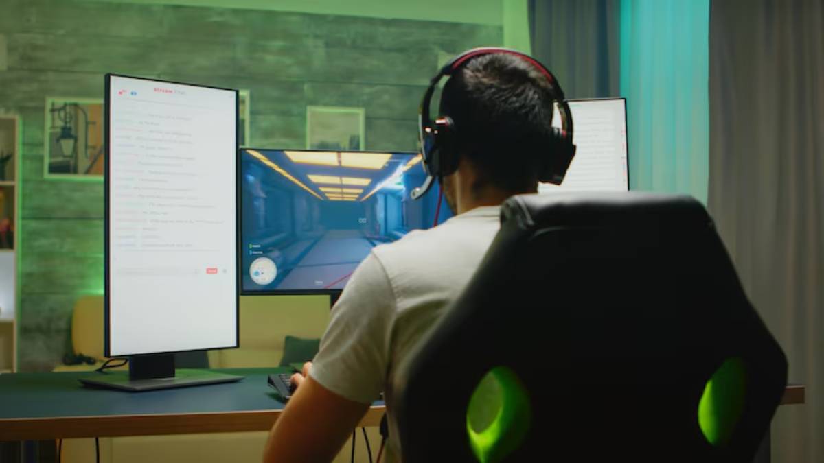 A gamer in a headset engages with dual monitors, one displaying gameplay and another showing a chat interface, in a cozy setting.