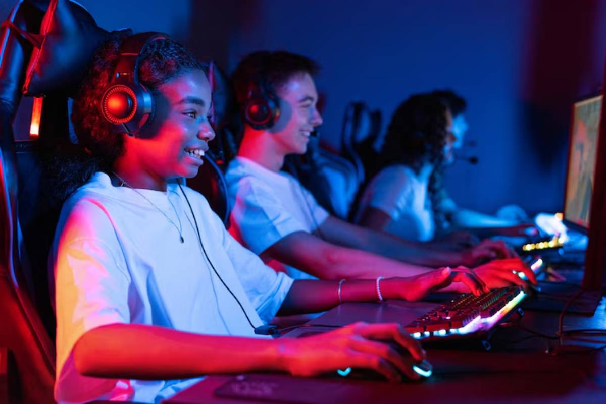 Three young gamers sit at illuminated desks, focused on their screens, wearing headsets and surrounded by a vibrant, colorful glow.