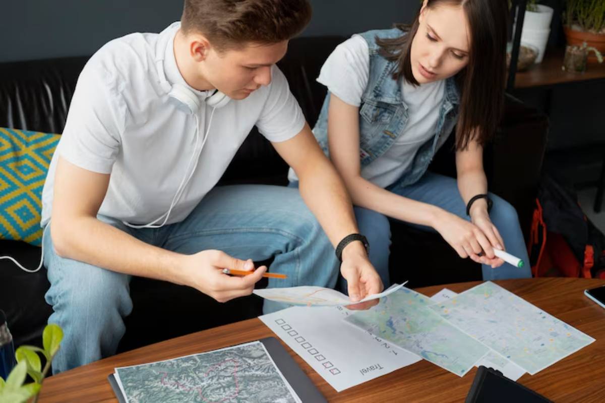 Two individuals are analyzing several maps on a table, discussing their plans with pens and notebooks at hand.