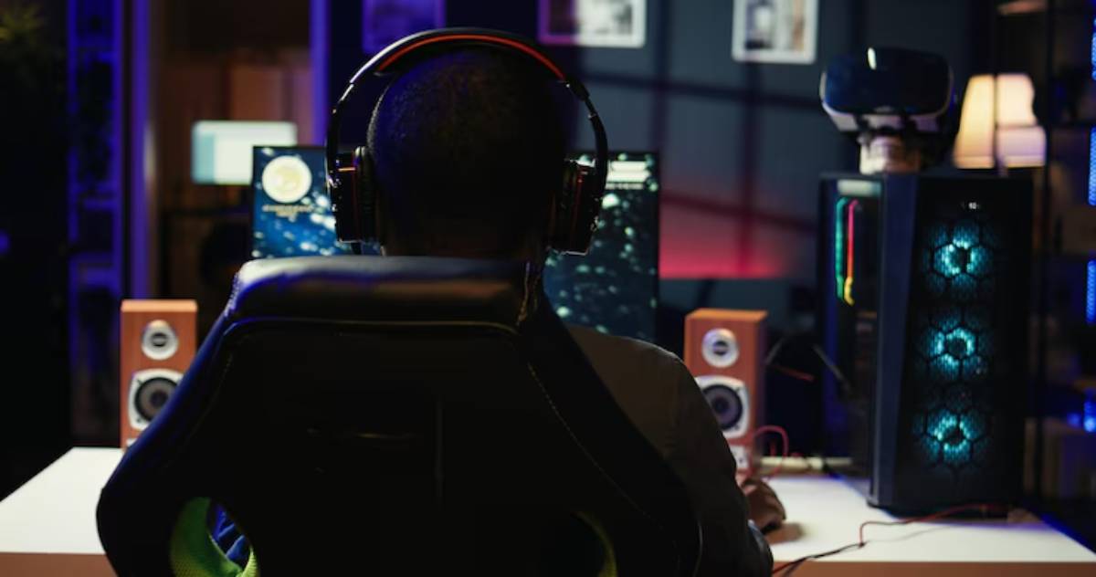 A gamer in a black headset sits in a stylish chair, focused on dual monitors displaying colorful graphics in a dimly lit room.