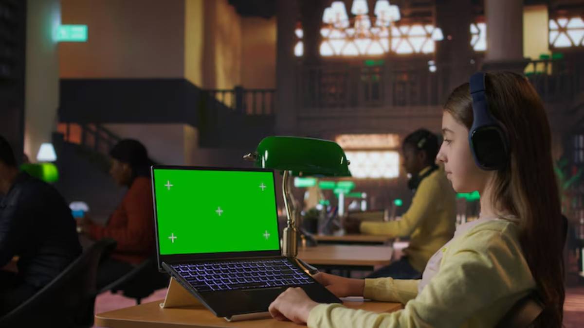 A person in a cozy library setting uses a laptop with a green screen, wearing headphones, while others work nearby.