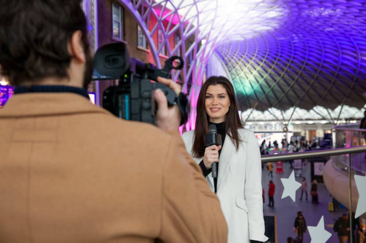 A woman in a white blazer holds a microphone while being filmed in a vibrant, modern train station with colorful lights and busy crowds.