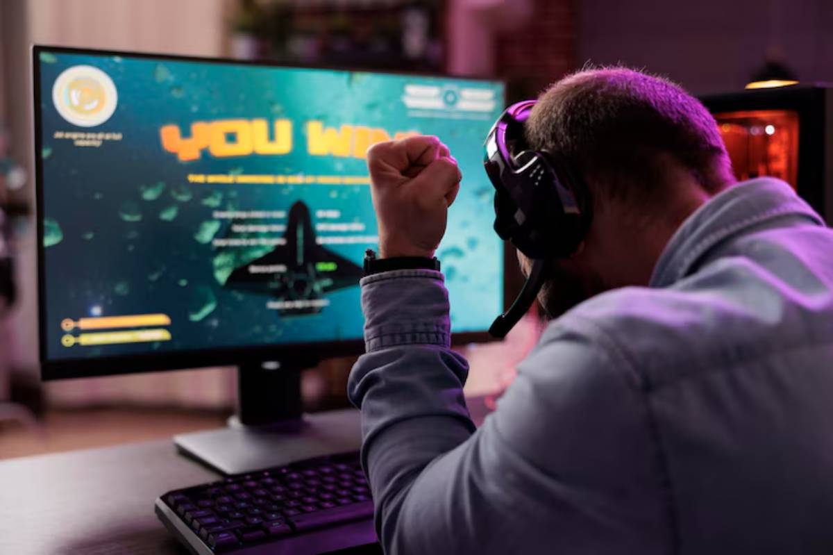 A gamer celebrates victory with a raised fist, wearing a headset, in front of a monitor displaying
