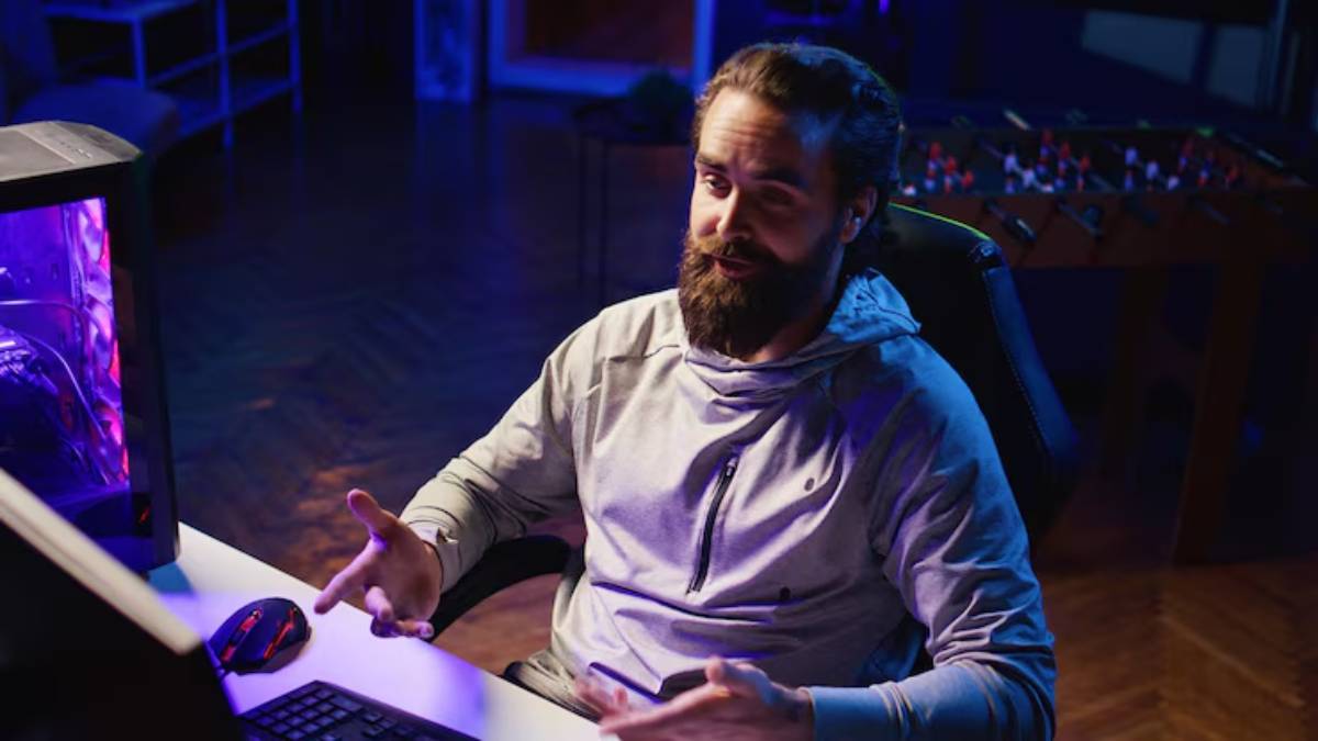 A person in a hoodie is gesturing while sitting at a desk with a gaming setup, surrounded by a dimly lit room with blue lighting.