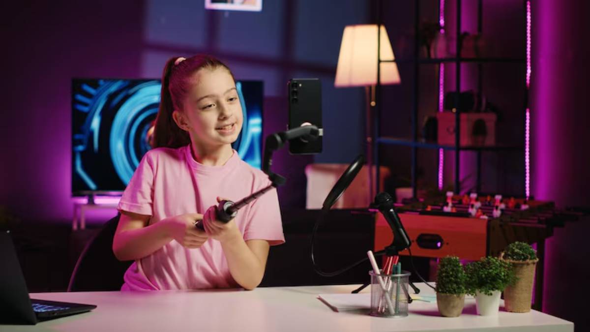 A person holds a selfie stick with a smartphone, surrounded by a laptop, microphone, and potted plants in a stylish, colorful room.