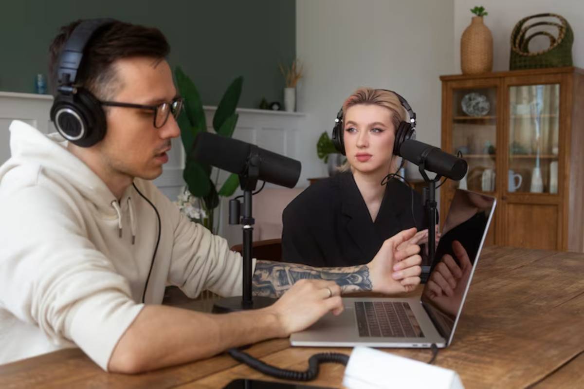 Two individuals with headphones and microphones sit at a wooden table, engaged in a podcast discussion, with a laptop in front of them.