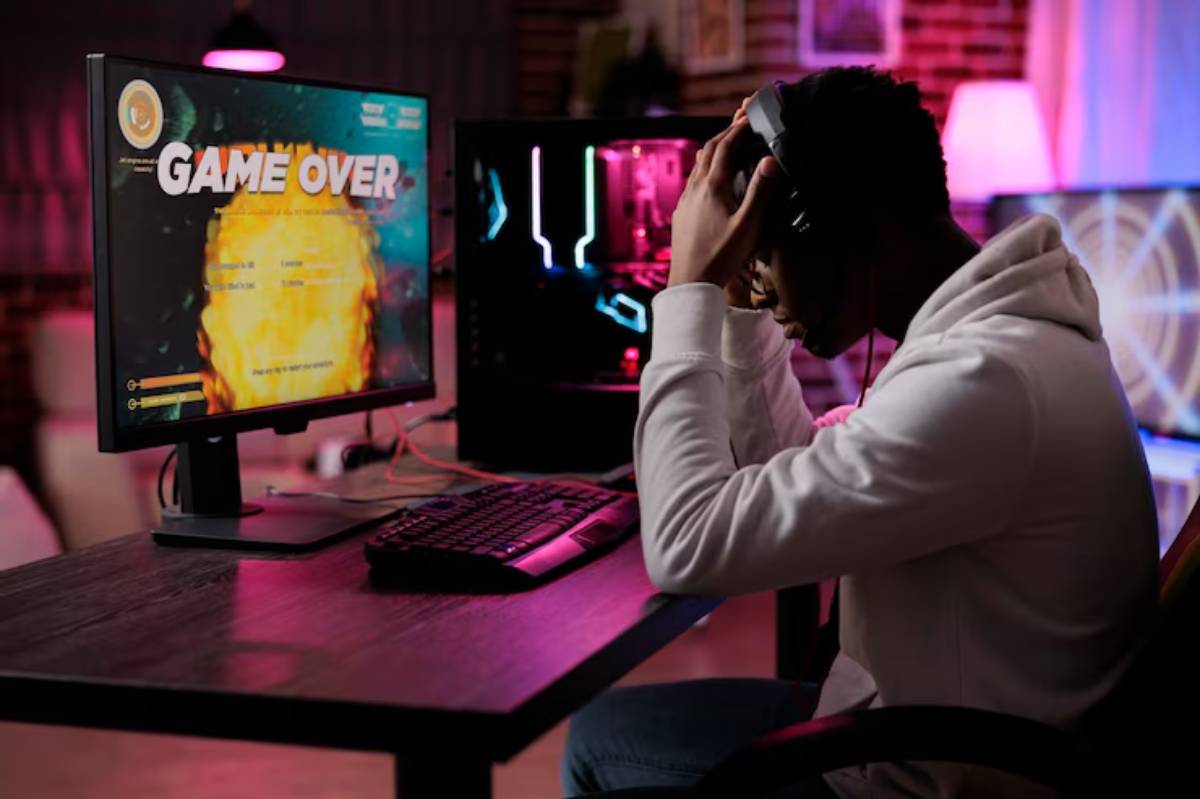 A gamer in a hoodie shows frustration after losing a game, staring at a monitor displaying 