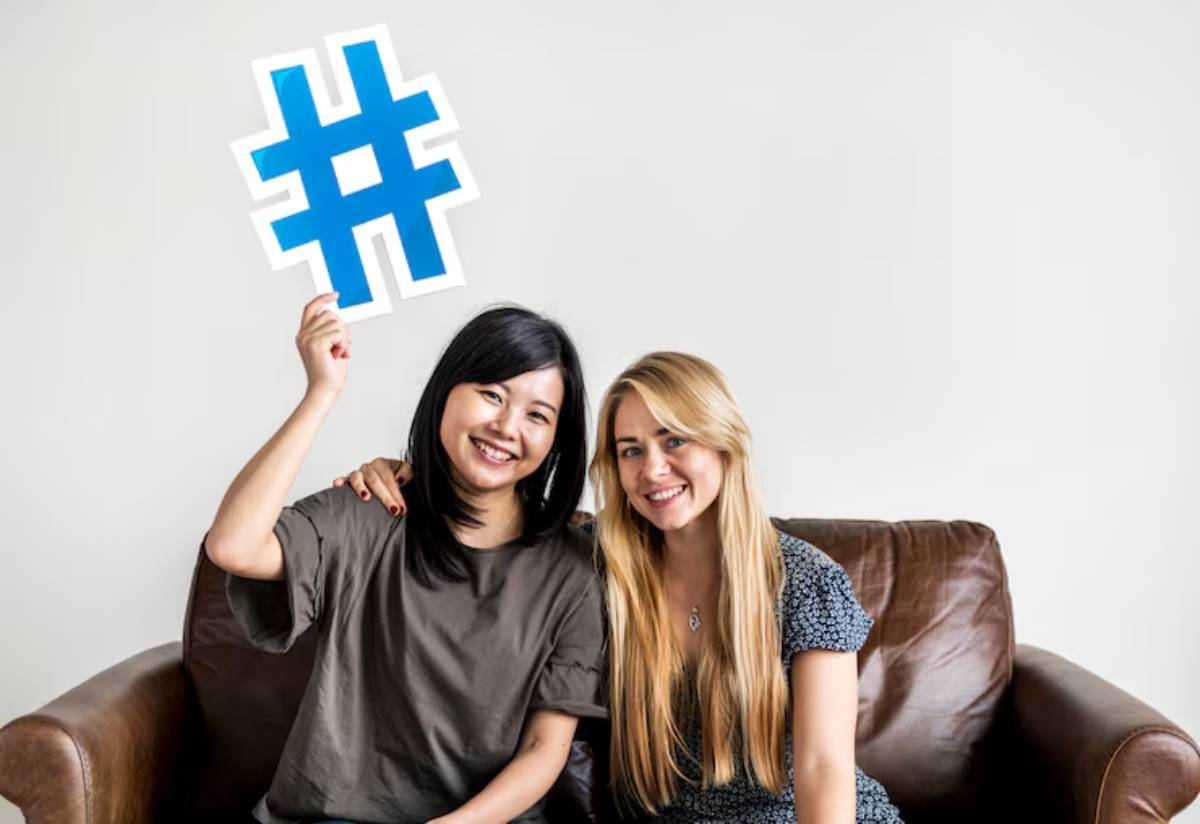 Two women sitting on a brown couch, one holding a large blue hashtag sign, smiling against a plain white wall.