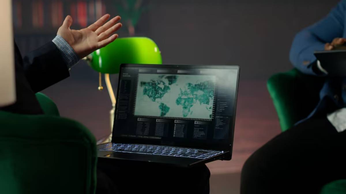  A person gestures while showing a laptop displaying a world map with data analytics, set in a cozy, dim-lit environment.