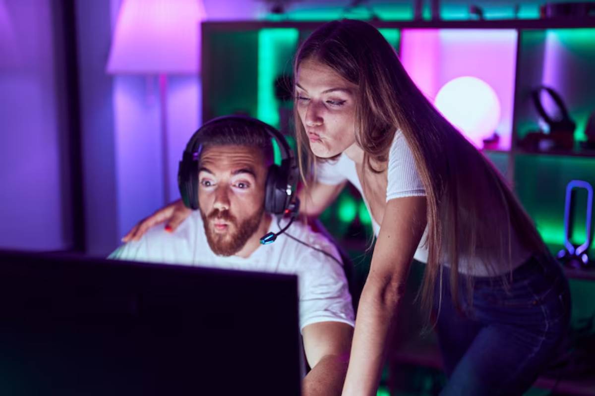 A woman leans over a man's shoulder in a colorful gaming setup, with ambient lighting creating a vibrant atmosphere.