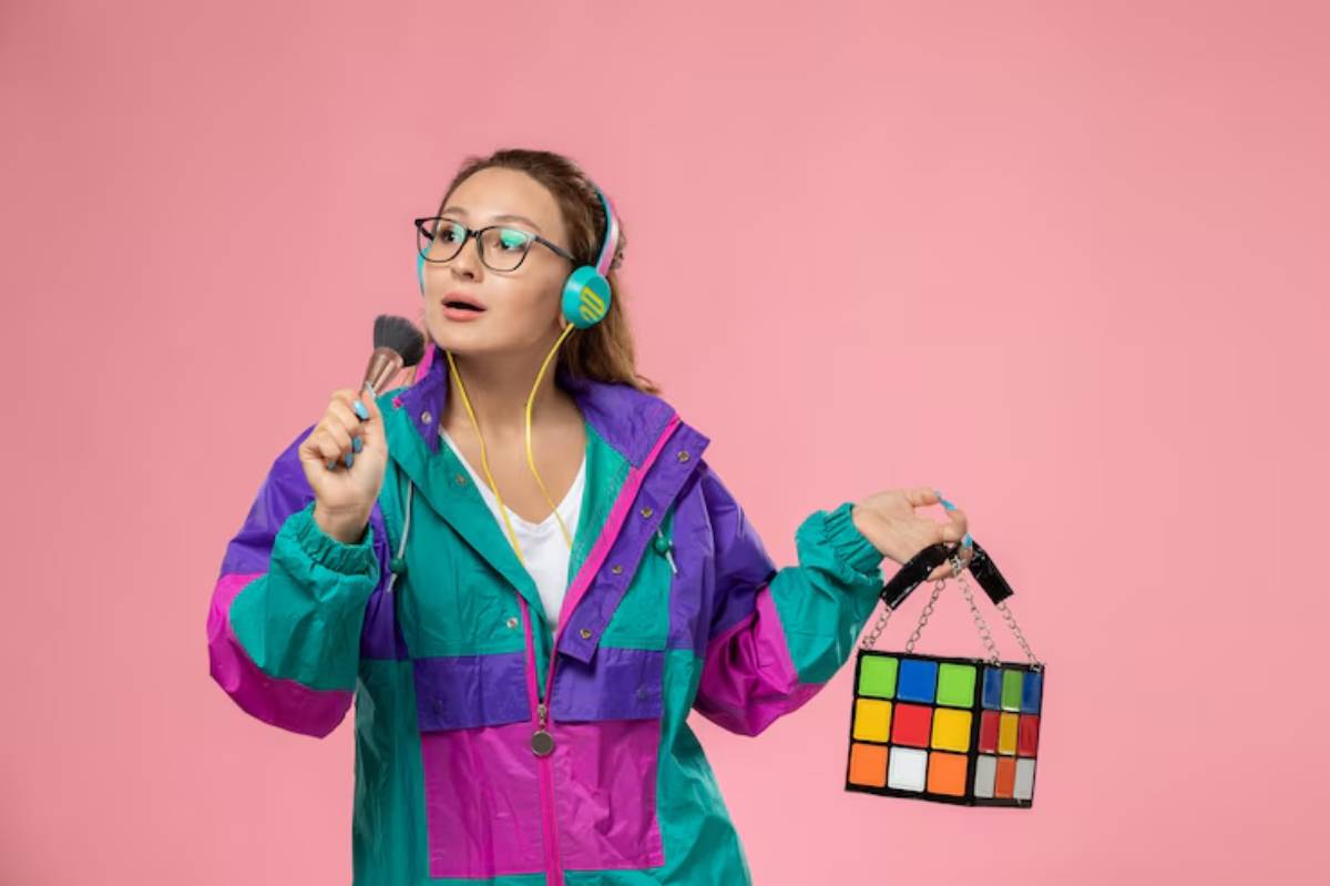 A person in a colorful 80s-style jacket holds a makeup brush in one hand and a Rubik's Cube-inspired handbag in the other against a pink background.