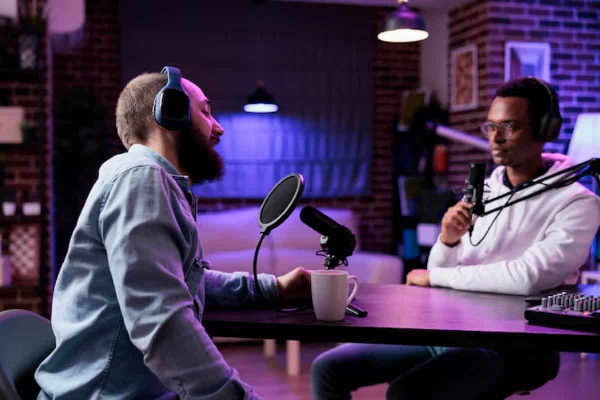Two individuals engage in a podcast conversation, seated at a modern table with microphones and headphones in a cozy studio setting.
